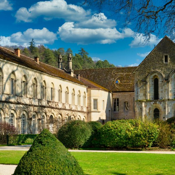 Vue extérieure d'une abbaye médiévale en pierre avec des jardins à la française aux arbustes taillés, sous un ciel bleu parsemé de nuages.